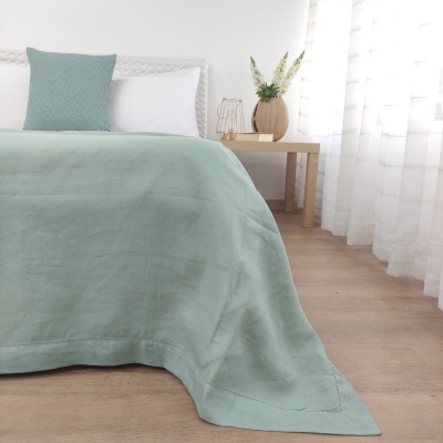 Cama com colcha verde claro e almofadas, ao lado mesa de madeira com vaso e janela com cortinas brancas