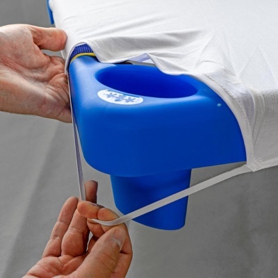 Mãos a esticar fita elástica branca para fixar capa numa mesa de massagem azul com orifício para rosto