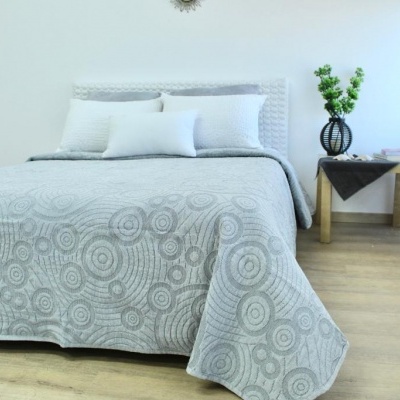 Cama com colcha cinzenta padrão círculos e almofadas brancas ao lado de mesa de cabeceira com vaso e flores.