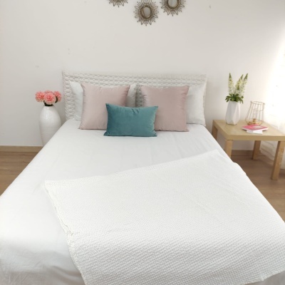Cama com almofadas cor de rosa e azul, cobertor branco dobrado, mesa com vaso e livros, parede branca com espelhos decorativos