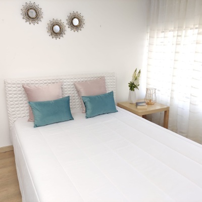 Cama branca com almofadas turquesa e rosa ao lado de mesa de madeira com decoração e espelhos redondos na parede.