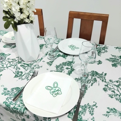 Mesa de jantar posta com toalha de mesa verde, pratos e guardanapos com desenho de coelho, copos transparentes, talheres e vaso branco com flores.