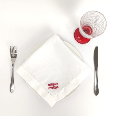 Mesa posta com prato branco, guardanapo branco bordado a vermelho, talheres em aço inox e copo com pé vermelho.