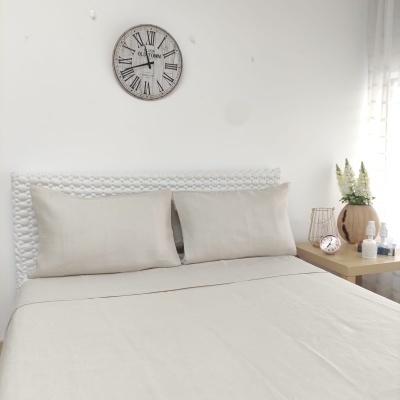 Cama com roupa cinza claro e cabeceira branca com relógio na parede e mesa ao lado