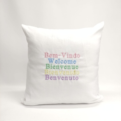 Almofada branca com bordado colorido de palavras de boas-vindas em vários idiomas