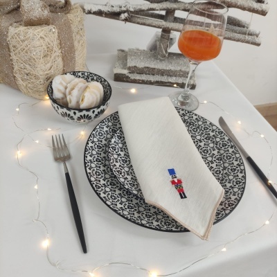 Mesa posta decorada com prato, guardanapo bordado, talheres pretos, tigela com pão e copa de vidro com bebida laranja.