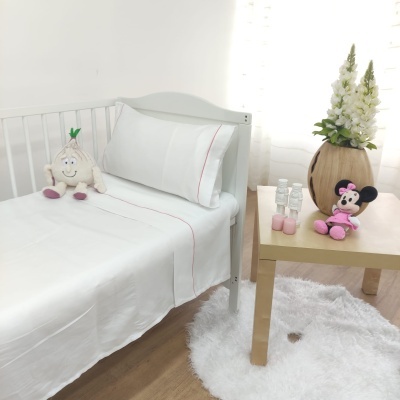 Cama de criança branca com lençóis e almofada brancos com risca vermelha e bonecos de peluche ao lado de mesa com vaso de flores
