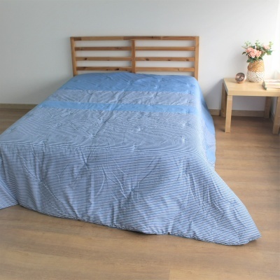 Cama de madeira clara com roupa de cama azul às riscas e mesa de cabeceira com vaso e relógio