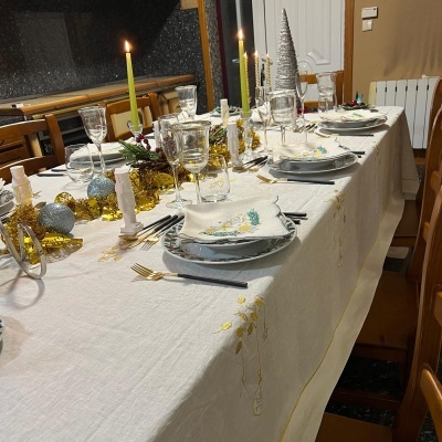 Mesa posta para festa com decoração natalícia de linho branco, velas, guardanapos decorados, talheres dourados e ornamentos festivos prateados e dourados.