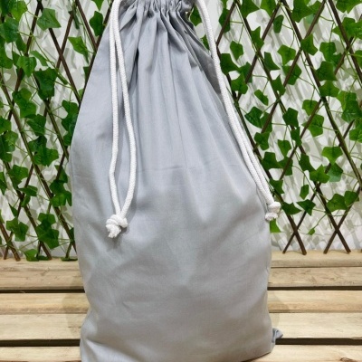 Saco cinzento de tecido com cordão branco numa superfície de madeira e fundo com folhas verdes