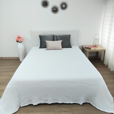 Cama com colcha branca e almofadas cinzentas e rosa numa divisória com decoração circular e um vaso de flores cor-de-rosa