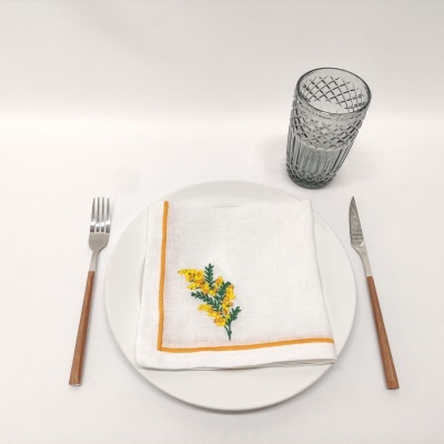 Conjunto de jantar minimalista com prato branco, guardanapo bordado, talheres com cabo de madeira e copo cinzento.