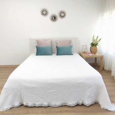 Cama de casal com colcha branca, almofadas rosa e azul, mesa de cabeceira com vaso de flores e três espelhos decorativos na parede