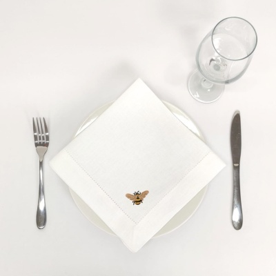 Guardanapo branco bordado com abelha sobre prato, talheres metálicos e copo de vinho