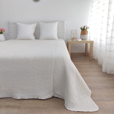 Colcha de cama branca texturizada com duas almofadas e mesa de cabeceira em ambiente iluminado