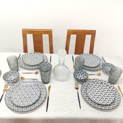 Mesa posta com conjunto de loiça preta e branca, copos cinzentos, garrafa de vidro e talheres dourados e pretos