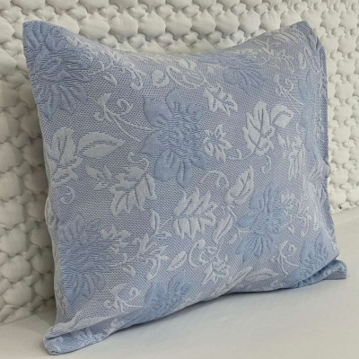 Almofada azul clara com padrão floral branco em relevo sobre colcha branca texturada