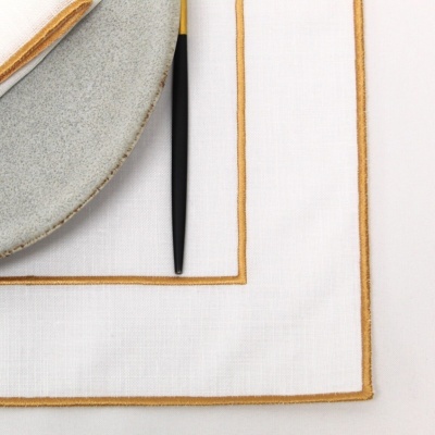 Guardanapos e toalha de mesa brancos com bordas douradas com um prato cinza e talher preto e dourado