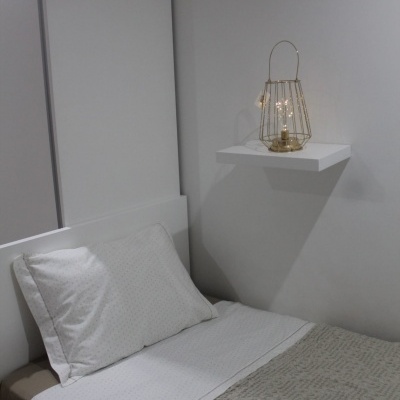 Cama branca com colchão e roupa de cama em tons neutros, prateleira com lanterna dourada na parede cinza