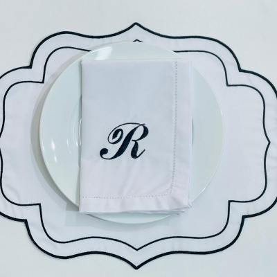 Prato branco com guardanapo branco bordado com a letra R em cima de um toalhete branco com bordas pretas onduladas