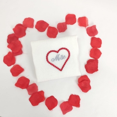 Toalha branca com bordado de coração vermelho e palavra Mãe, rodeada por pétalas de rosa vermelha formando um coração