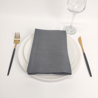 Mesa posta com prato branco, guardanapo cinzento dobrado, talheres em preto e dourado e copo de vidro.