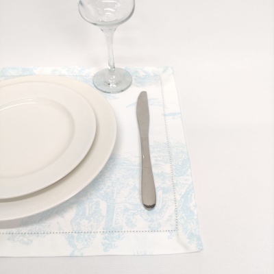 Jogo americano branco com padrão azul claro, pratos brancos, faca metálica e taça de vidro numa mesa branca