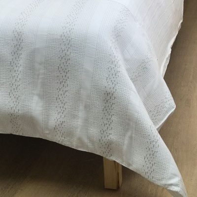 Capa de edredão branca com padrão de riscas verticais e textura, sobre cama em ambiente com chão de madeira clara