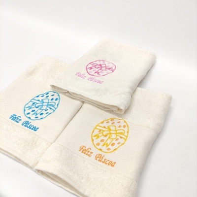 Três toalhas brancas bordadas com texto 'Feliz Páscoa' em rosa, azul e amarelo.