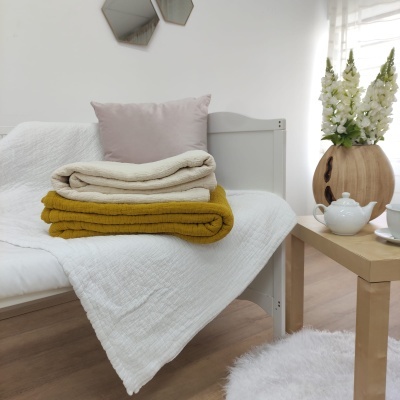 Cama branca com colcha e mantas empilhadas ao lado de mesa com bule e jarra com flores