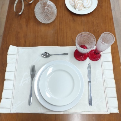 Conjunto de loiça branca com talheres e copos de vidro sobre placemat bege e mesa de madeira.