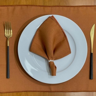 Conjunto de mesa com guardanapo e base castanho-avermelhado, prato branco, talheres com cabos pretos e lâminas douradas.
