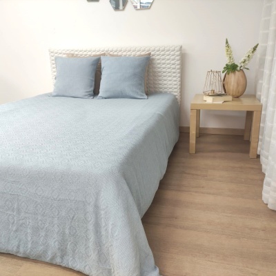 Cama de casal com colcha azul clara e duas almofadas azuis ao lado de mesa de madeira com vasos decorativos
