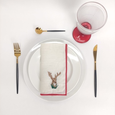 Conjunto de mesa com prato branco, guardanapo com bordado de rena, talheres pretos e dourados, e copo com base vermelha
