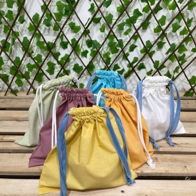 Seis sacos de tecido multicoloridos com cordão sobre paletes de madeira com treliça e folhas ao fundo