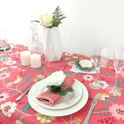 Mesa posta com toalha rosa floral, pratos brancos, guardanapos rosa com flores brancas, copos de vidro, garrafa de cristal e vela.