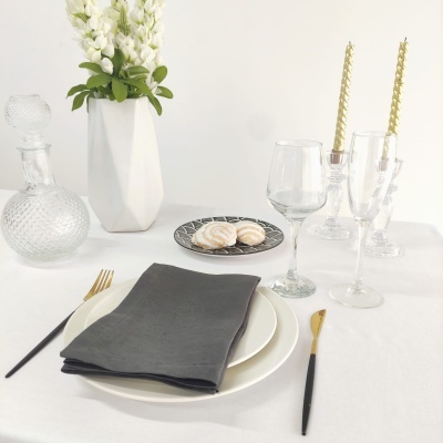 Mesa posta elegante com prato branco, guardanapo cinza, talheres preto e dourado, copo de vinho, copos de licor, doces em prato preto, jarro de vidro e vaso geométrico com flores brancas.