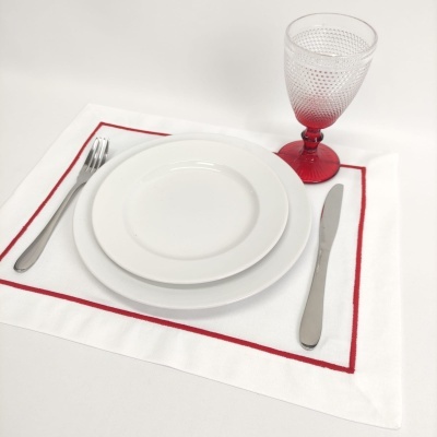 Conjunto de pratos brancos, talheres metálicos, guardanapo branco com borda vermelha e copo de vidro vermelho texturizado