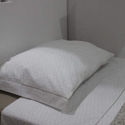 Travesseiro com fronhas brancas com padrão de pequenos pontos e detalhe bordado sobre lençol branco com padrão igual numa cama