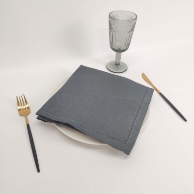 Mesa posta com guardanapo cinzento, prato branco, talheres pretos e dourados e copo cinzento de vidro.