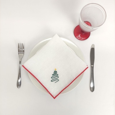 Mesa posta com guardanapo branco com borda vermelha e árvore de Natal verde sobre prato branco, talheres metálicos e copo com base vermelha