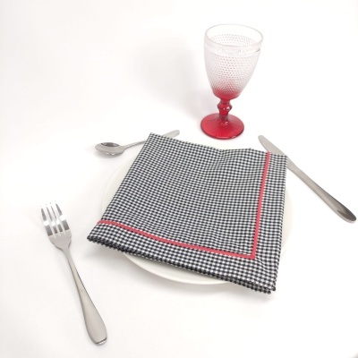 Conjunto de jantar com prato branco, guardanapo quadriculado preto e branco com linha vermelha, talheres em aço inoxidável e copo de vidro vermelho com padrão de bolinhas.