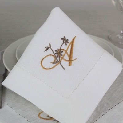 Guardanapo branco bordado com letra A dourada e castanha em prato branco com talheres e tecido cinza