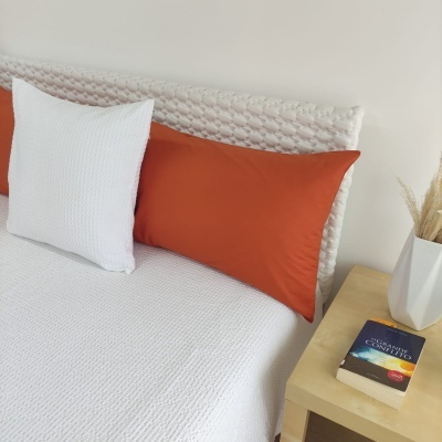 Cama com roupa de cama branca e almofadas laranja e branca junto a mesa de cabeceira com livro e vaso branco.