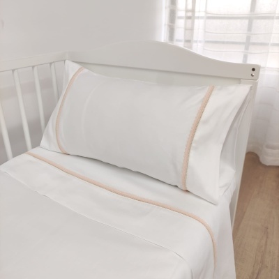 Lençol e fronha brancos com linhas cor de rosa e renda numa cama branca junto a uma janela com cortinas transparentes