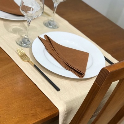 Mesa posta com prato branco, guardanapo castanho, garfo preto e dourado, copo transparente, toalha bege e mesa madeira clara