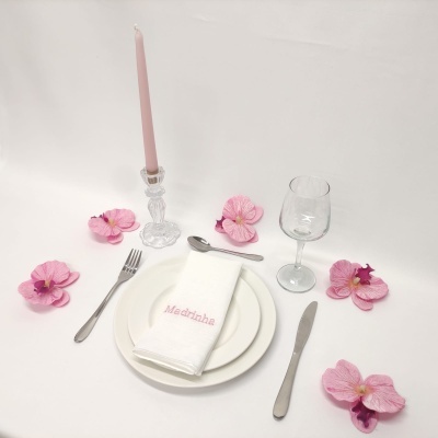 Conjunto de mesa com talheres de metal, pratos brancos, guardanapo branco bordado, copo de vidro, castiçal com vela rosa e flores rosa