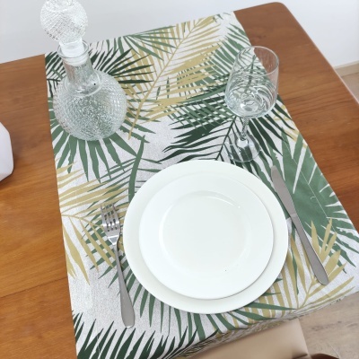 Mesa posta com toalha branca estampada com folhas verdes e douradas, prato branco, talheres, copo e garrafa de vidro