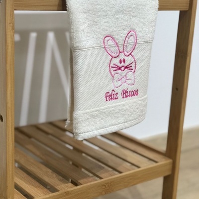 Toalha branca com bordado de coelho e texto Feliz Páscoa sobre estrutura de madeira