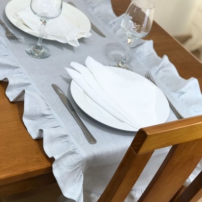 Mesa posta com toalha cinza clara com bordas franzidas, pratos e guardanapos brancos, talheres e copos de vidro.